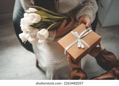 Đôi tay của đứa trẻ cầm một hộp quà xinh đẹp có ruy băng và hoa tulip trắng. Nhìn từ trên xuống, cận cảnh. Chúc mừng ngày của mẹ.