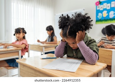 Estudiante negra afroamericana haciendo un examen en la escuela primaria. Adorables niñas sentadas en el interior de la mesa, sintiéndose molestas y deprimidas mientras aprenden con el maestro en el jardín de infantes.