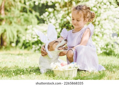 Bambina carina che abbraccia il suo cagnolino indossando un costume con le orecchie da coniglio pasquale. Cestino con uova decorate e fiori.