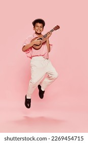Djali i vogël afrikan i lumtur emocional, me rroba të stilit retro dhe pozon me kapele me kitarë ukulele të izoluar në sfond rozë. Koncepti i muzikës, fëmijërisë, edukimit, modës dhe aspiratës