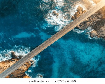 Aerial view of bridge, sea with waves and stones at sunset in Lofoten Islands, Norway. Landscape with beautiful road, transparent blue water, rocks. Top view from drone of highway in summer. Transport