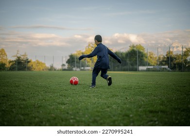 Copil care joacă fotbal. Un băiat asiatic dă cu piciorul în fotbalul roșu.