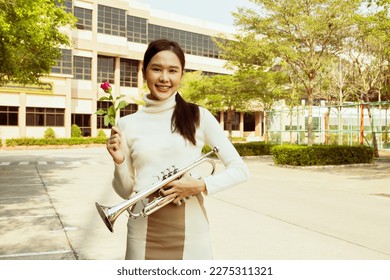 International music teacher in school : Portrait beautiful healthy asian teacher holding bright red rose and trumpet in front of school building Show flowers with friendly smiles to lovely students.