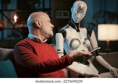 Idősebb férfi és női android robot együtt tévézik otthon éjjel, ő tartja a távirányítót