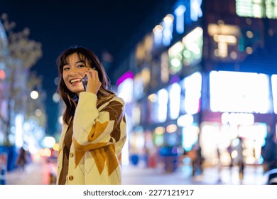 Young Asian woman talking on mobile phone during travel in city at night. Attractive girl enjoy and fun modern outdoor lifestyle nightlife city street using portable device with wireless technology.