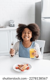 Gadis Afrika-Amerika yang bahagia memegang garpu dan segelas jus jeruk di dekat pancake sambil menikmati sarapan