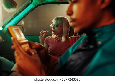 Um casal retrô estiloso está sentado em um carro estacionado. O homem está jogando um videogame, e sua namorada está ao lado dele com um pirulito.