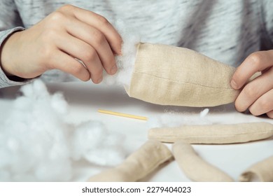 알아볼 수 없는 여성 디자이너 손이 부드러운 장난감을 바느질하는 클로즈업. 발과 신체 부위를 만들고, 테이블에 면으로 채웠다. 어린이 선물 가게, 매장. 소규모 사업, 창의적인 직업 및 튜토리얼