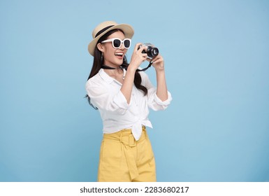 Glücklich lächelnde junge asiatische Touristin im Sommerhut, die mit der Kamera steht und ein Foto isoliert auf blauem Studiohintergrund macht