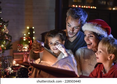 Una familia feliz encuentra una tableta digital en un regalo en la noche de Navidad, cerca de la estufa de leña y el árbol de Navidad iluminado en su sala de estar, la madre usa un gorro de Papá Noel