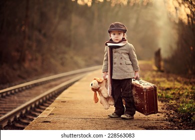 Adorable niño en una estación de tren, esperando el tren con maleta y oso de peluche
