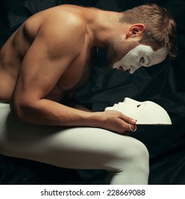 Koncepti i dyfishtë i misterit. Portret artistik i interpretuesit të cirkut me geta duke mbajtur dhe duke parë maskën veneciane (volto bianco), duke pozuar mbi pëlhurë të zezë. Trup muskuloz dhe nxirje perfekte. E shtënë në studio