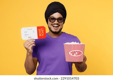 Sretni mladi Sikh Indijac nosi kravate svoj tradicionalni turban dastar ljubičastu majicu u 3D naočalama gleda film film drži u ruci kantu s kartom kokica u kinu izolirano na žutoj pozadini studio