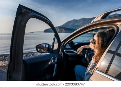 Junge glückliche Frau genießt den Sonnenuntergang im Auto an der Küste sitzend