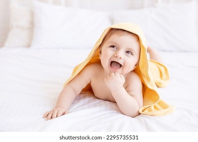 carino bambino biondo con un asciugamano giallo in testa dopo un bagno o un bagno su un letto bianco a casa in una stanza luminosa sorride, il concetto di cura e igiene dei bambini