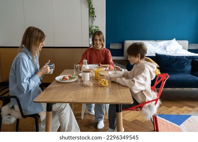 母親と2人の子供、10代の女の子と男の子が、自宅のテーブルに座って一緒に朝食を食べています。ワンルームマンションのモダンで居心地の良いインテリア。健康的な栄養、母性、育児のコンセプト。