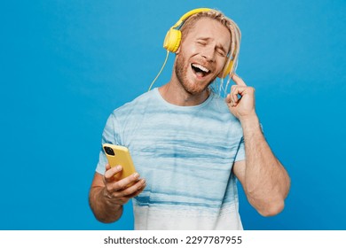Young blond man with dreadlocks 20s he wear white t-shirt headphones listen to music hold in hand use mobile cell phone isolated on plain pastel light blue background studio People lifestyle concept