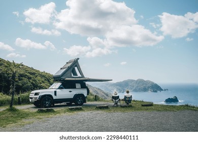 Couple sitting in a camping chair in nature with a view of Island in a beautiful blue sky with an overlanding car with a rooftop tent and an awning.