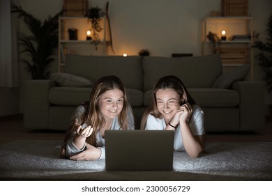 Two young women lying on the floor having video call on laptop at night. Sisters watching social media content in the living room.