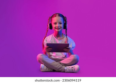 Digital Native, Generation Alpha Concept. Дете пред тийнейджърска възраст, щастливо, красиво и очарователно момиче в лятно облекло, седнало на пода над футуристичен фон, използващо безжични слушалки и цифров таблет, пространство за копиране