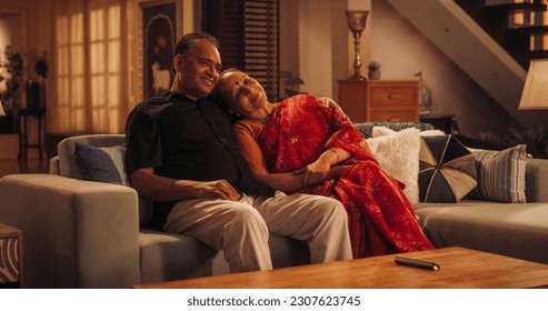 Waktu Bersama yang Menyenangkan: Pasangan Lansia India Menikmati Malam yang Santai di Rumah, Menonton Televisi, Berpelukan, Tersenyum. Wanita Cantik Menyandarkan Kepalanya di Dada Suaminya. Cinta Sejati Selama Bertahun-tahun
