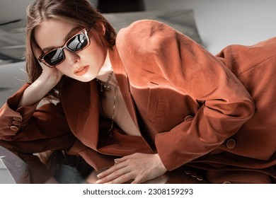 junge Frau trägt Terrakotta und trendigen Anzug mit Blazer und goldener Halskette und posiert mit modischer Sonnenbrille auf grauem verspiegeltem Hintergrund, schönes Modell, Spiegelung, faszinierend