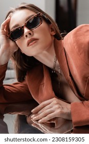 junge Frau trägt Terrakotta und trendigen Anzug mit Blazer und goldener Halskette und posiert mit modischer Sonnenbrille auf grauem Spiegelhintergrund, schönes Modell, Spiegelbild, Blick in die Kamera