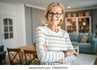 Chân dung một phụ nữ da trắng tóc vàng trưởng thành đeo kính ở nhà, mỉm cười hạnh phúc nhìn vào máy ảnh, tự tin mặc áo len trong phòng sáng, không gian sao chép