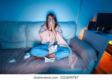Femme malade assise sur un canapé recouvert d'une couverture tard dans la nuit, souffrant de grippe et de fièvre, se mouchant avec des mouchoirs en papier tout en regardant la télévision