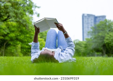 African American woman is lying down in the grass lawn inside the public park holding book in her hand during summer for reading and education