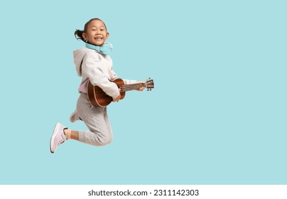 Vajzë shkolle, nxënëse e lumtur aziatike nxënëse e shkollës që kërcen nga gëzimi me dorë që mban ukulele me kitarë, portret me trup të plotë i izoluar në sfond të thjeshtë pastel blu të çelët