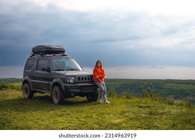 A happy girl in a bright orange sweater, is standing next to a small SUV with a roof rack. View point on the top of the mountain, Dniester Subich river, Ukraine. Copy space