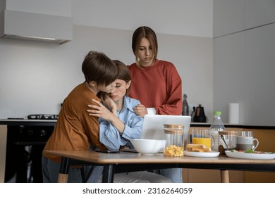 母親はオンラインで仕事中に問題を抱えています。十代の子供たちはストレスの多い瞬間に母親を支えました。男の子と女の子が、自宅のキッチンのテーブルに座ってノートパソコンで作業している母親を抱きしめています。家庭と仕事を両立しています。