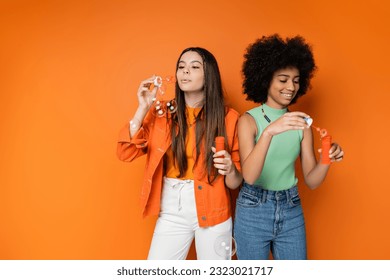 Stylish brunette teenage girl blowing soap bubbles near cheerful african american girlfriend with bold makeup while standing together on orange background, fashionable and trendy clothes