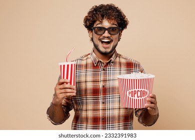 Mladi veseo zabavan sretan Indijac u 3D naočalama gleda filmski film držeći kantu kokica šalicu soda pop kole gazirana voda u kinu izoliran na običnoj pastelnoj svijetlo bež pozadini studijski portret