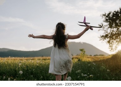 Vue arrière d'une petite fille enjouée qui court avec un avion sur fond de montagnes étonnantes pendant le coucher du soleil chaud de l'été. Concept de liberté de rêve. L'enfant court sur le terrain en tenant dans ses mains un avion jouet