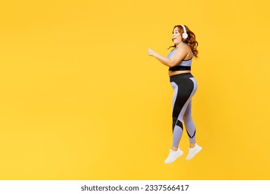 Full body young plus size fat fit woman wear blue top warm up train listen music in headphones jump high do aerobic exercise isolated on plain yellow background studio home gym. Workout sport concept