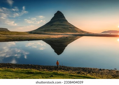 Aerial view of majestic symmetry volcano Kirkjufell mountain with lake reflection and male tourist standing during sunrise morning in summer at Snaefellsnes peninsula, Iceland
