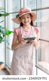 美しいピンクのよだれかけドレスと花柄の帽子をかぶったアジアの若い元気な女の子が、タッチスクリーンのタブレット コンピューターを使ってインターネットを閲覧したり、自宅で一人でオンライン ストリーミング ゲームをしたりしながら笑顔で立っています。