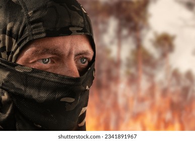 Military man in a camouflage mask against the backdrop of a burning forest. The concept of military action. Man at war. Fire after explosions. Forest fire. Arsonist. Closeup of the face.