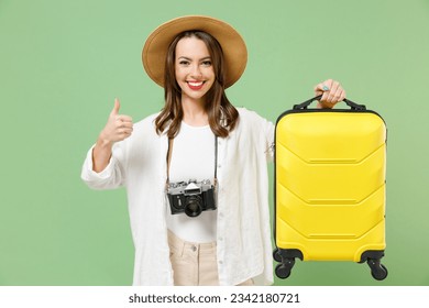 Traveler tourist woman in casual clothes hat camera hold yellow suitcase valise show thumb up gesture isolated on green background Passenger travel abroad weekends getaway Air flight journey concept