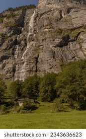 Blockhütte im Sommertag mit steilen Bergklippen dahinter und einem langen Wasserfall in Lauterbrunnen, Schweiz