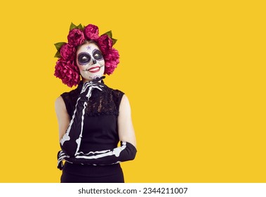 Retrato de una mujer feliz con maquillaje de calavera de azúcar, guantes de esqueleto y flores en la cabeza, sonriendo aislada sobre un fondo amarillo con una expresión facial pensativa. Concepto de Halloween y Día de los Muertos.