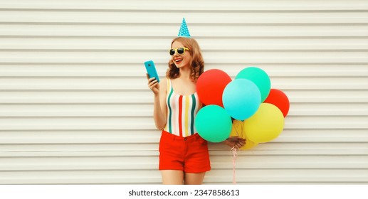 Happy cheerful young woman in festive birthday hat celebrating with smartphone and bunch of colorful balloons having fun wearing striped t-shirt, sunglasses on white background