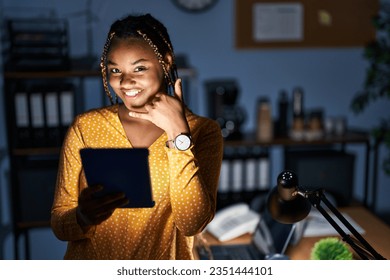 African american woman with braids working at the office at night with tablet smiling doing phone gesture with hand and fingers like talking on the telephone. communicating concepts.