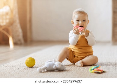 Bayi balita pirang yang lucu dan ceria bermain dengan mainan anak-anak di rumah sambil duduk di lantai karpet di ruang tamu. Potret anak kecil yang lucu dan tersenyum menggunakan mainan warna-warni, teether kunyah, ruang fotokopi