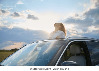 Ein schönes junges Mädchen schaut aus der Luke eines Autos und genießt den Sonnenuntergang und die Sonnenstrahlen, die auf ihrem Gesicht glitzern. Eine schöne Frau sonnt sich im Sonnenuntergang. Autoreisekonzept.