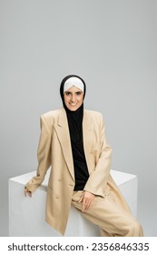 moderna muslimanka poslovna žena sa blistavim osmijehom i gleda u kameru u blizini bijele kocke na sivom