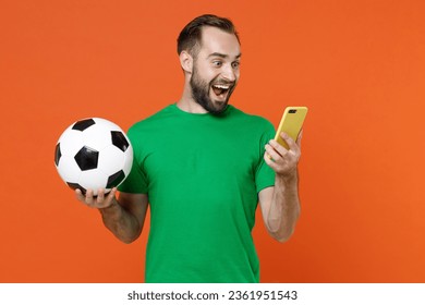 Un joven aficionado al fútbol sorprendido con una camiseta verde anima a su equipo favorito con un balón de fútbol usando un teléfono móvil aislado sobre un fondo naranja. Concepto de estilo de vida, ocio y deporte para personas