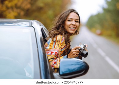Stylish female tourist films nature from a car window with a retro camera and enjoys the autumn weather. Adventure, vacation concept. Active weekend.